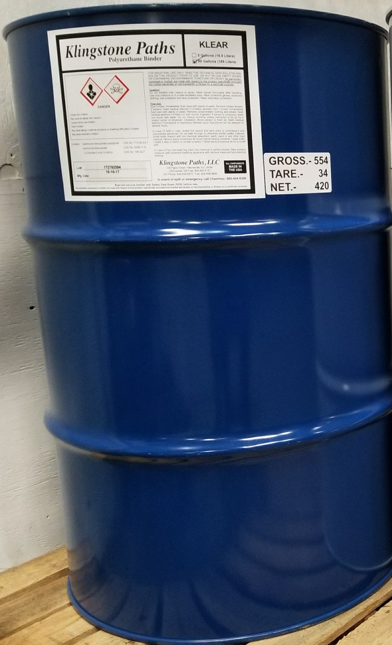 A large blue metal drum barrel with a white product label reading 'Klingstone Paths' and 'KLEAR', photographed indoors against a light wall on a wooden surface.